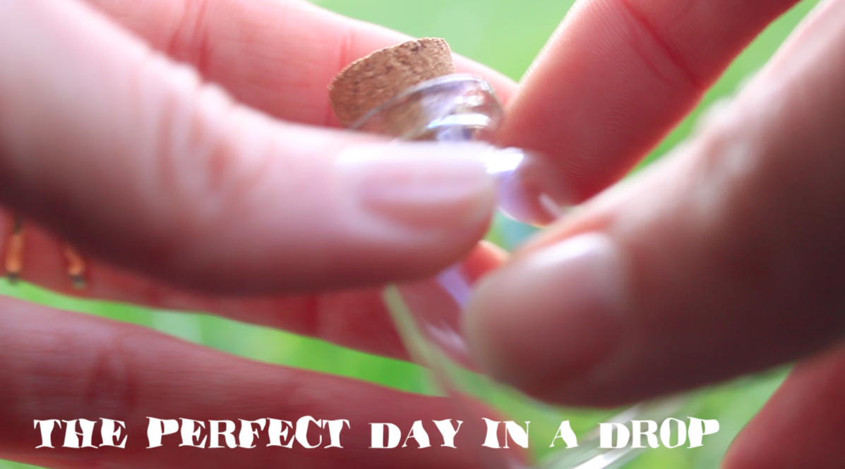 Right and left hands' fingers holding a tiny glass jar. In the background a green field. On the left corner of the image, a white text in waterish bold font says : The Perfect Day in a Drop.